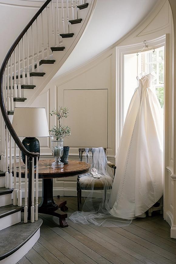 Wedding dress hanging by a curved staircase, with veil on a chair, bridal shoes, and greenery on a round table in window light