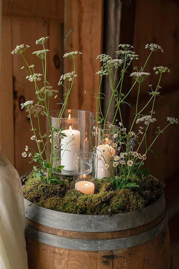 Wedding centerpiece with candle centerpiece in glass cylinder vases, wildflowers and greenery on a wooden barrel against a rustic wooden wall