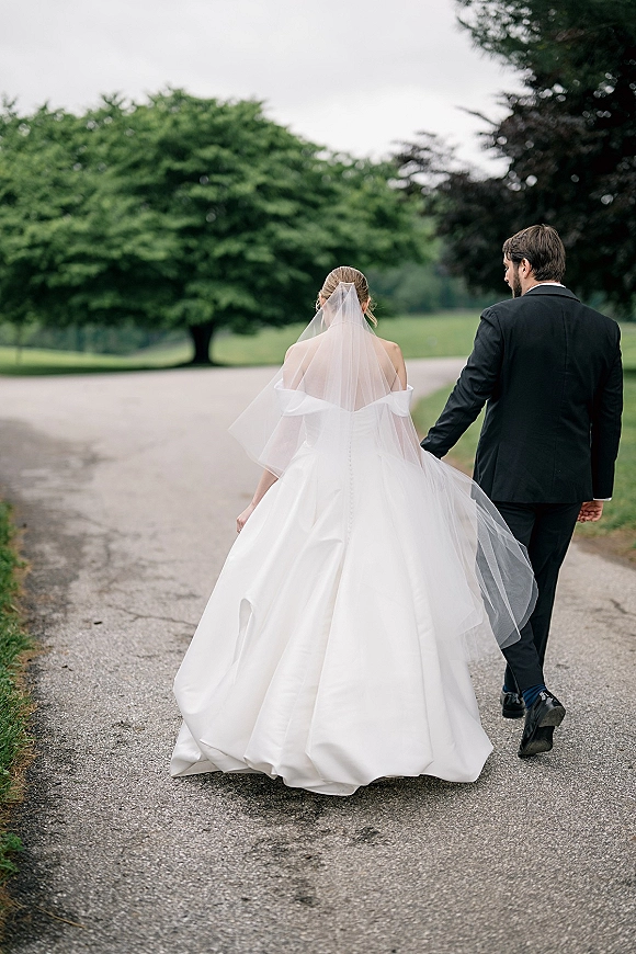 Couple portrait of bride and groom walking away hand in hand, her cathedral veil and buttoned train flowing on a tree-lined road