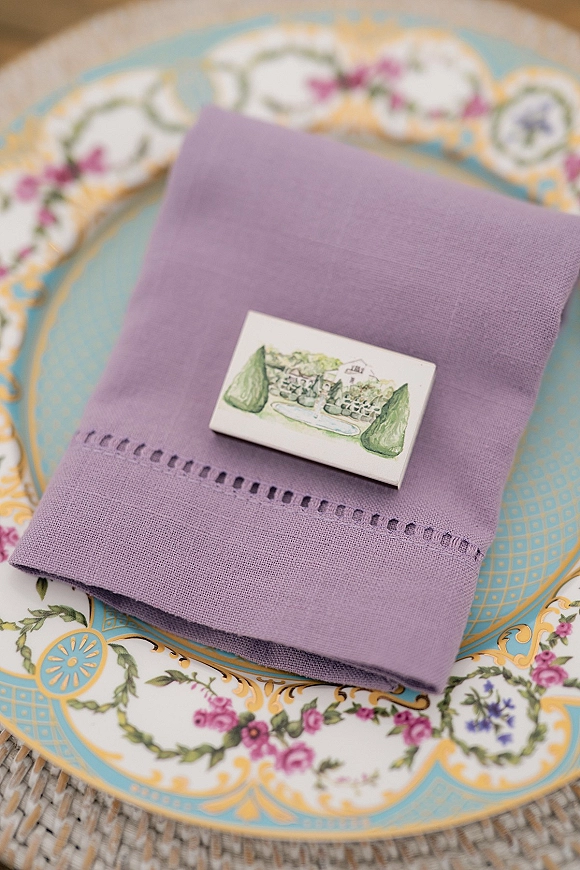 Wedding place setting with a lavender napkin and illustrated place card, layered floral china and blue patterned plates on a woven charger tabletop
