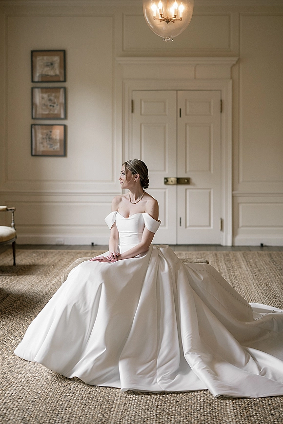 Bridal portrait of a seated bride in an off the shoulder wedding dress with a pearl necklace, on a bench in a white paneled room with chandelier