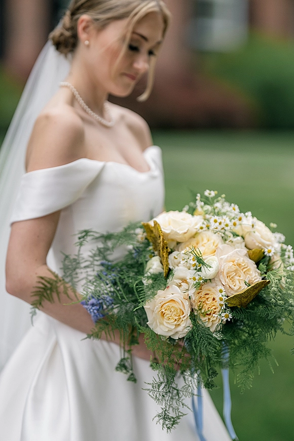 Bridal portrait of a bride holding bouquet in an off the shoulder wedding dress with veil, pearl necklace, and roses on an outdoor lawn