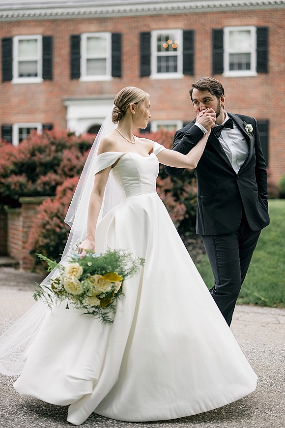 Couple portrait of bride and groom walking as he kisses her hand, her veil and bouquet flowing on a garden walkway by a brick building