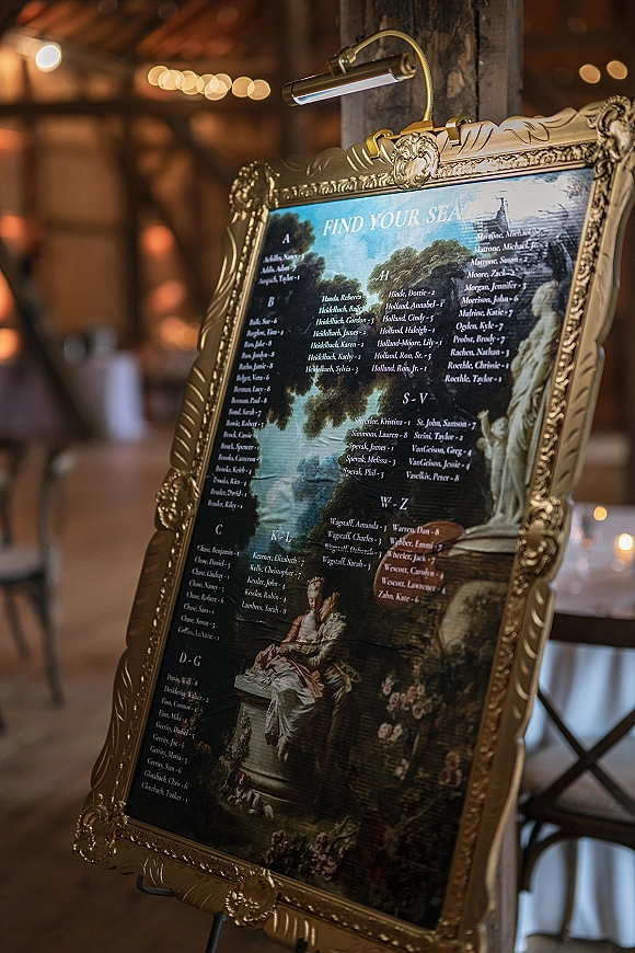 Wedding seating chart in an ornate gold frame on an easel, lit by a picture light in a rustic reception room with string lights