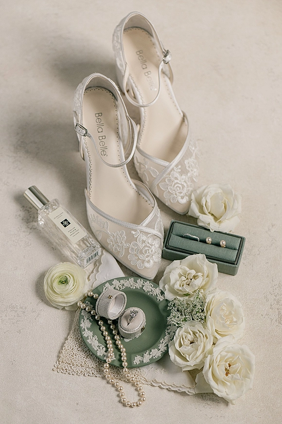 Bridal details flat lay with lace wedding heels, pearl jewelry, rings in boxes, perfume bottle, and white roses on a neutral surface