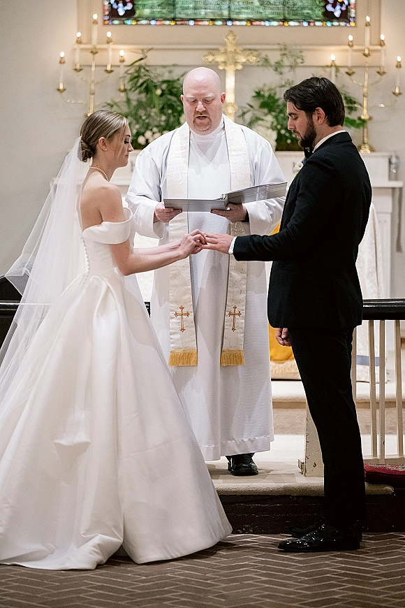 Wedding ceremony moment as bride places a ring on the groom’s hand at a church wedding ceremony, altar cross and stained glass behind