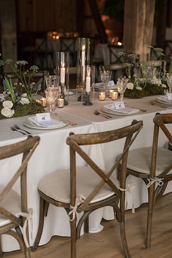 Reception tablescape with a candlelit wedding table featuring taper candles in glass cylinders, moss runner, white linens, and rustic barn beams behind