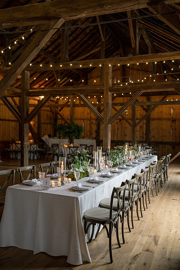 Reception tablescape with a long banquet table wedding setup, taper candles and greenery garland on white linens under barn string lights