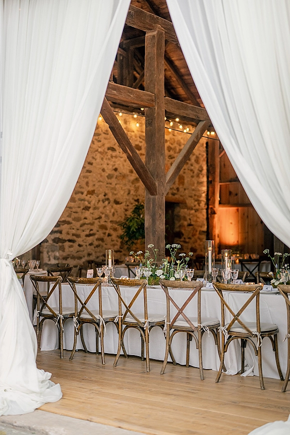Reception tablescape with taper candles and bud vases on farm tables, white draping and string lights in a rustic barn with stone wall