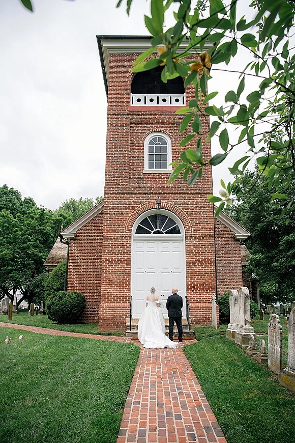 Couple portrait of bride and groom from behind walking away, veil and dress train flowing on a brick walkway to white church doors