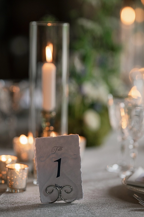 Reception tablescape with wedding table number card beside pillar candle in glass hurricane, low floral centerpiece, and soft bokeh lights behind