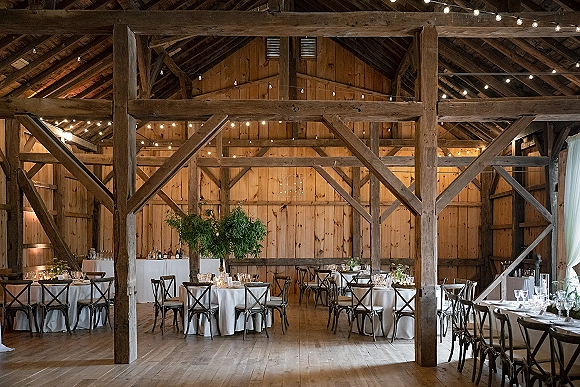 Barn reception decor with rustic barn reception string lights over round tables, white linens, cross-back chairs, and greenery centerpieces in a wood-beam barn interior