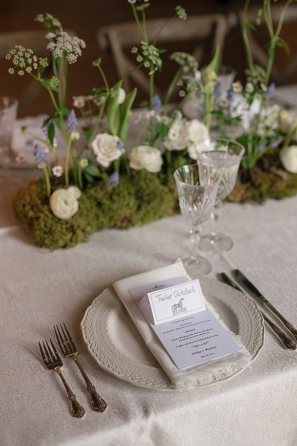 Reception tablescape with a wedding place setting, moss runner and wildflower centerpiece, menu and place cards on ivory linen in indoor reception space