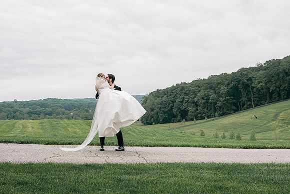 Wedding kiss portrait of groom lifting bride as her long veil and dress train flow, set against rolling hills and an overcast sky
