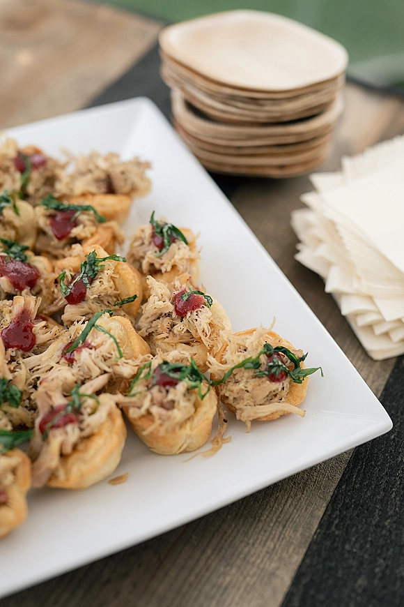 Wedding appetizers on a white serving platter, mini pastry cups topped with shredded meat and herb garnish, red sauce drizzle on a wood table outdoors