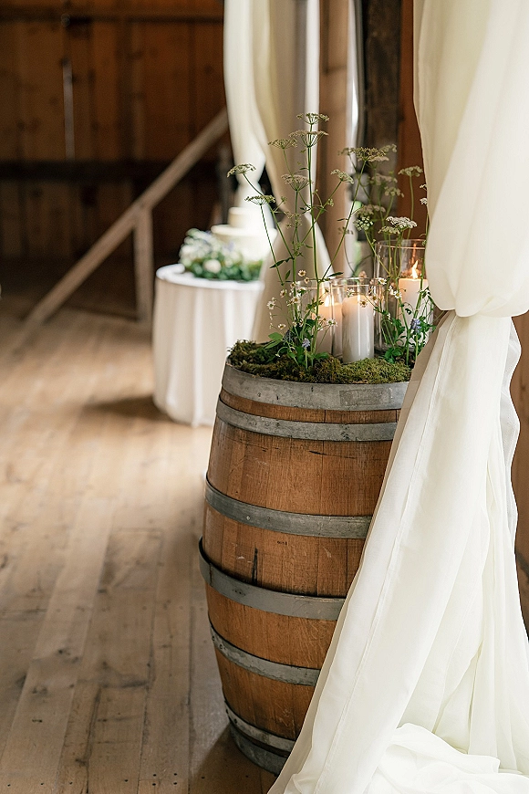 Wedding aisle decor with barrel wedding aisle decor featuring moss, wildflowers, greenery, and hurricane vases with pillar candles in a rustic barn setting