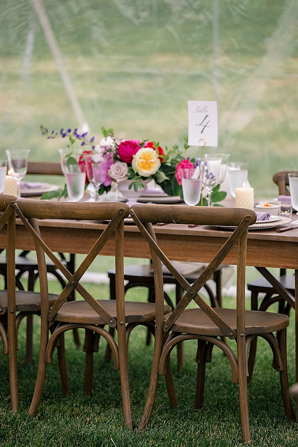 Reception tablescape on a wood farm table wedding with cross back chairs, floral centerpiece, candles, and glassware under a clear tent on a lawn