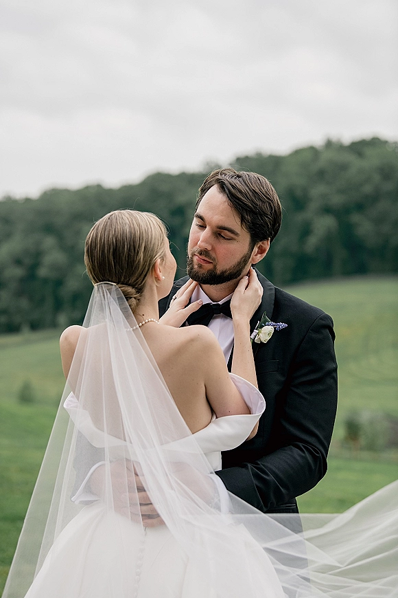 Couple portrait of bride and groom embrace as she touches his face, veil blowing in wind across a green field under cloudy sky