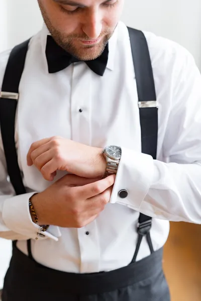 Groom getting ready, adjusting watch in a white dress shirt with black bow tie and suspenders against a clean white wall indoors
