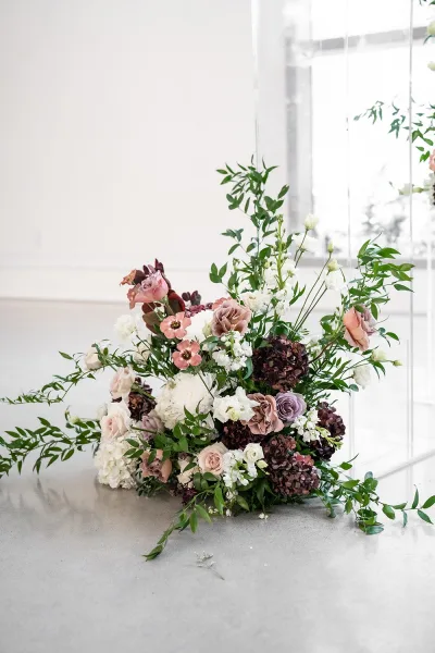 Floral arrangement of wedding ceremony flowers with roses, hydrangea, and greenery beneath a hanging acrylic panel by a white wall window