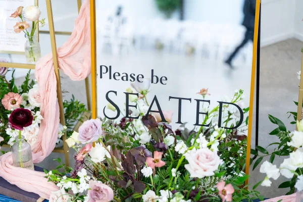 Wedding signage please be seated sign in a gold frame with blush draping and rose greenery florals on a white floor with blurred chairs behind