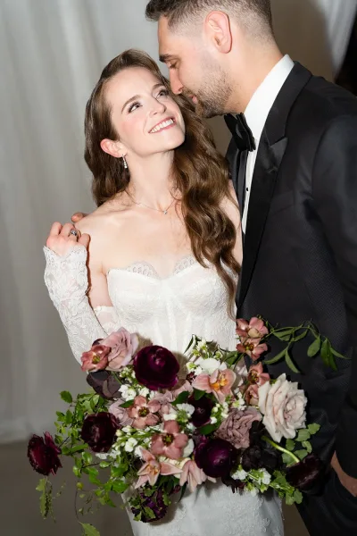 Couple portrait of bride looking at groom as he embraces her, holding a burgundy and blush bouquet before a white draped backdrop