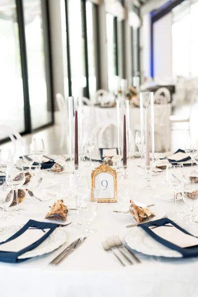 Reception tablescape with navy and white wedding table settings, taper candles in glass hurricanes, and gold vintage table numbers by tall windows