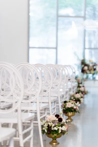 Ceremony aisle decor with wedding aisle flowers in gold urns beside white chairs, set on a polished floor by large windows overlooking greenery