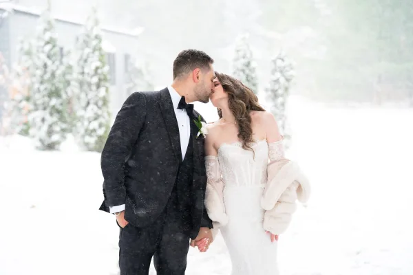 Wedding kiss portrait of bride and groom kissing in falling snow, she wears a faux fur wrap and lace sleeves beside evergreens and a house
