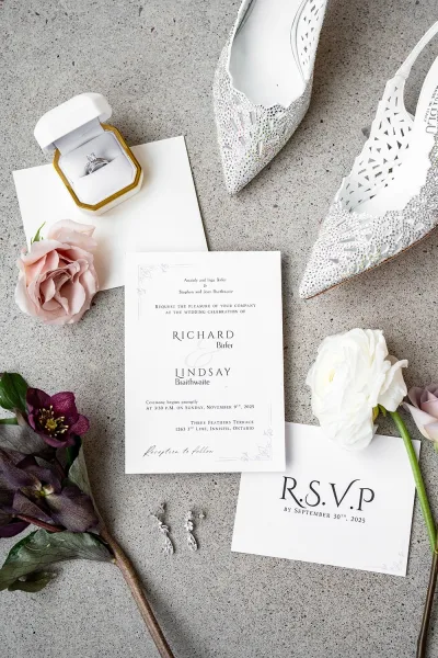 Invitation suite wedding invitation flat lay arranged with RSVP card, envelope, ring box, bridal shoes, earrings and blooms on stone surface
