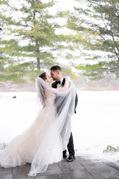 Wedding couple portrait of bride and groom embrace under her veil, nose to nose in snow on an outdoor patio with evergreens behind