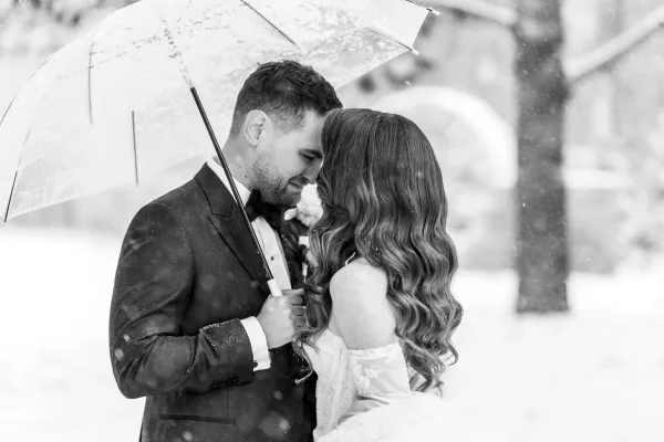 Couple portrait with a clear umbrella as bride in off-the-shoulder lace dress and groom in tuxedo embrace under falling snow by trees