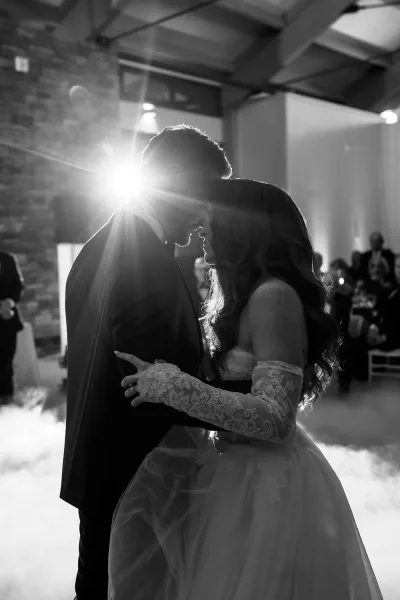 First dance as bride in a strapless gown and lace gloves holds her groom on foggy reception floor, backlit in brick hall with guests