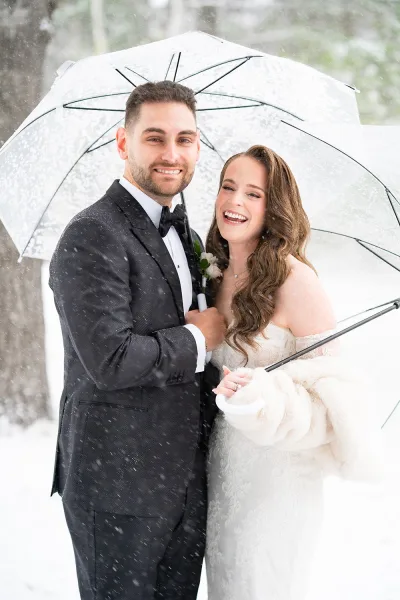 Couple portrait of a winter bride and groom under a clear umbrella as snow falls, her fur wrap and his tuxedo set against snowy trees