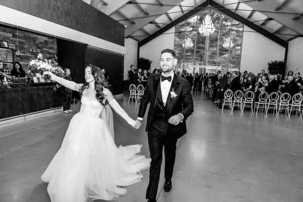 Recessional moment as bride and groom entrance holding hands, bride raising bouquet under chandeliers in a brick-walled reception hall