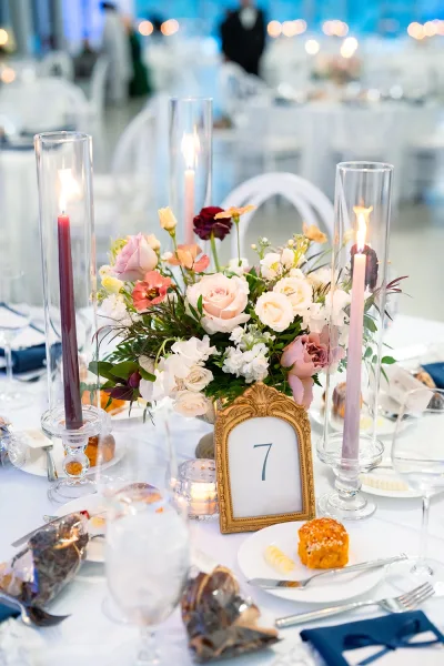 Reception tablescape with wedding table centerpiece of roses and greenery, taper candles in glass holders, navy napkins on white linens under soft lighting