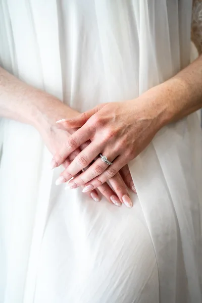 Engagement ring with oval diamond sparkle on a French-manicured hand, paired with a wedding band against soft white tulle skirt fabric