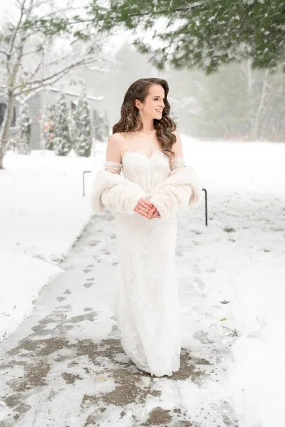 Bridal portrait of a bride in a strapless lace gown with faux fur wrap, smiling on a snowy path among winter trees near a house