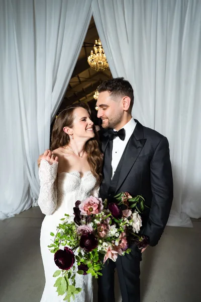 Couple portrait of bride in a strapless lace wedding dress and groom in tuxedo holding a bouquet beneath draped curtains and chandelier