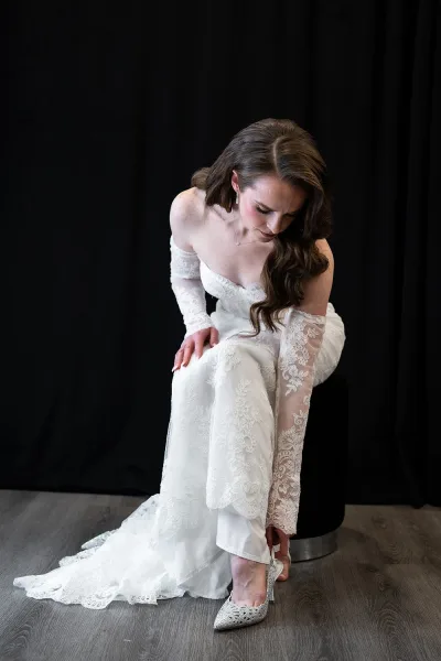 Bride portrait of a seated bride putting on shoes, wearing an off-the-shoulder lace wedding dress and jewelry against a black backdrop
