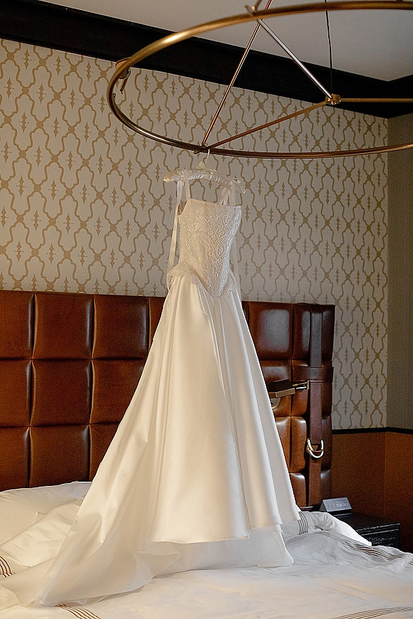 Wedding dress with beaded bodice hanging on a hanger, satin skirt and ribbon straps in a hotel room above a made bed