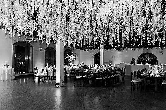 Reception tablescape with hanging floral ceiling, candlelit centerpieces, and mixed round and banquet tables in an indoor ballroom with arched windows