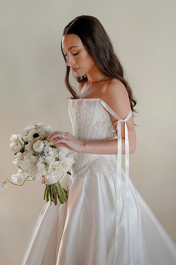 Bridal portrait of a bride holding bouquet in side profile, wearing a strapless satin ball gown with ribbon ties on a neutral studio backdrop