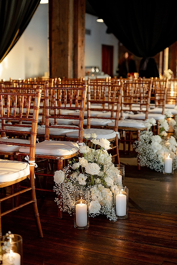 Ceremony aisle decor with aisle floral arrangements of white roses and baby's breath, lined with pillar candles in glass vases on wood floors