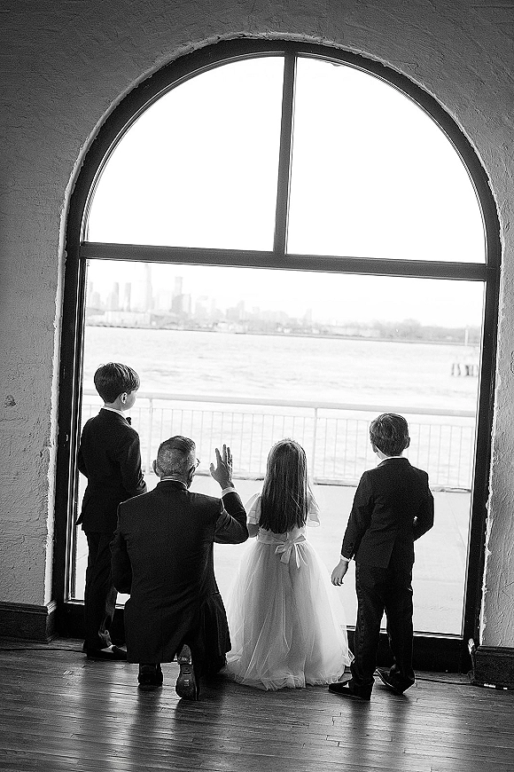 Wedding kids portrait of a flower girl and ring bearer in tuxedos, seen from behind in window light at an arched window with city skyline