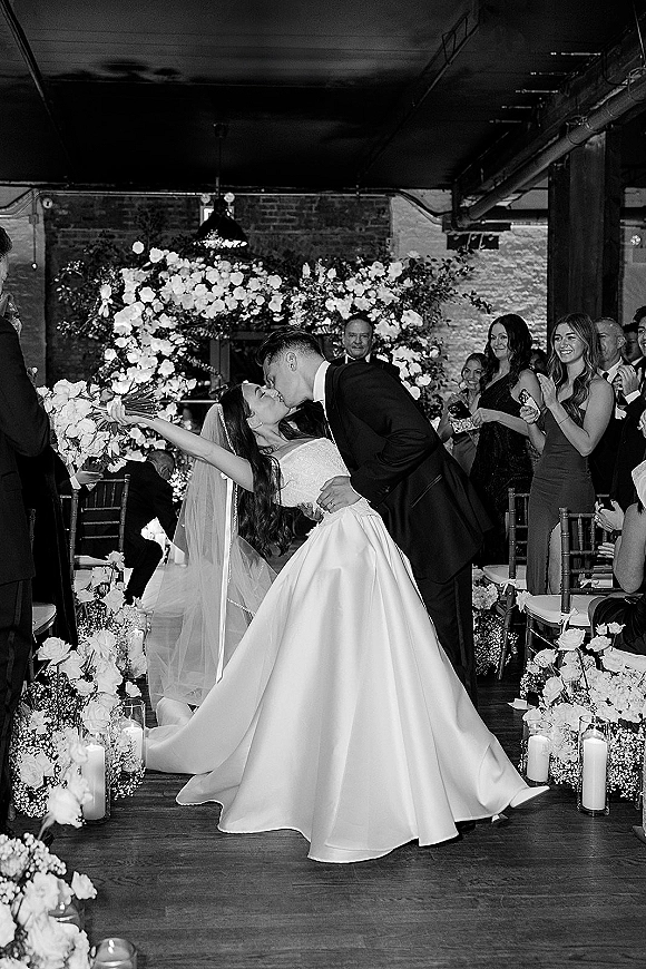 Wedding kiss portrait of bride and groom in a dip kiss wedding photo, bouquet raised, under a floral arch with candles and brick wall backdrop