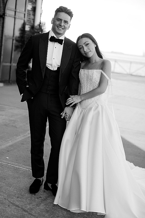 Couple portrait in a black and white wedding portrait style, bride in strapless gown leaning on tuxedo groom on an outdoor walkway by railing