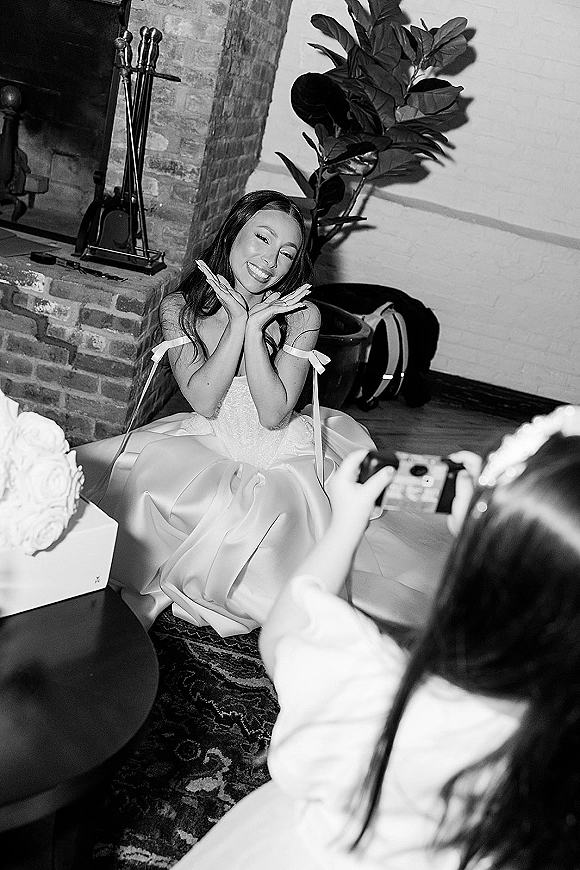 Bridal portrait of a smiling bride sitting on the hardwood floor in a satin ribbon-strap gown, holding a bouquet by a brick fireplace
