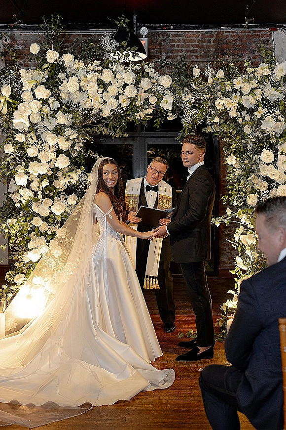 Ceremony moment as couple exchanging vows, holding hands beneath a white rose floral arch in an indoor brick-walled venue with candles