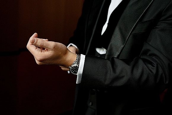 Groom detail as he adjusts his cuff beside a wristwatch, wearing a black tuxedo with bow tie and pocket square against a dark wall